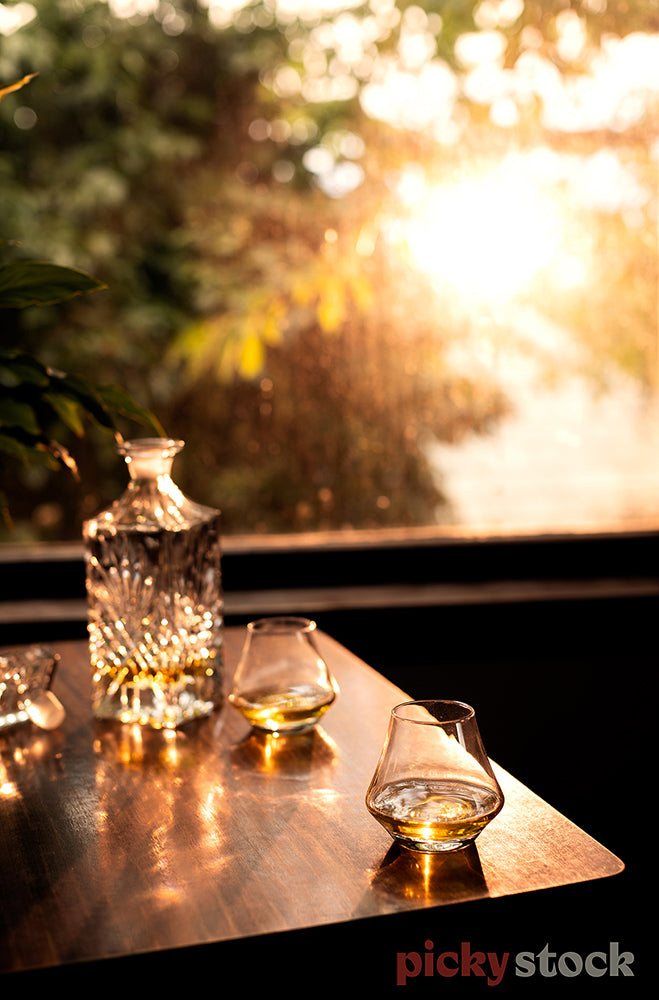 Three glasses and large crystal decanter sitting on top of wooden table. View to from outside window of large tree and ocean in the distance. 