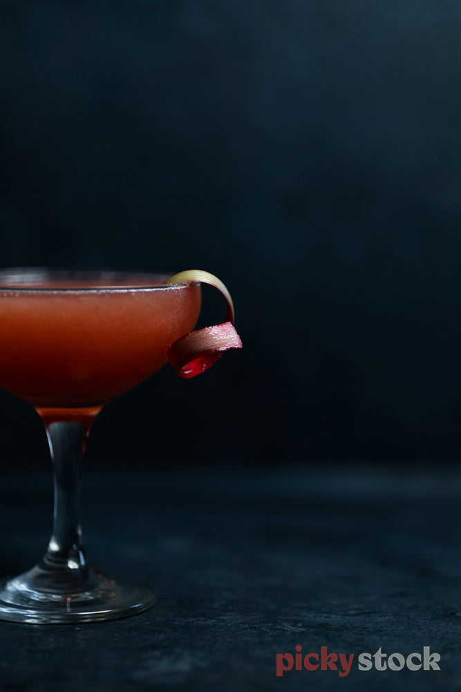 Close up shot of cocktail siting on dark surface with dark background. Cocktail is bright with a red colour filed to the top. A piece of rhubarb is curled on the side of the glass. 