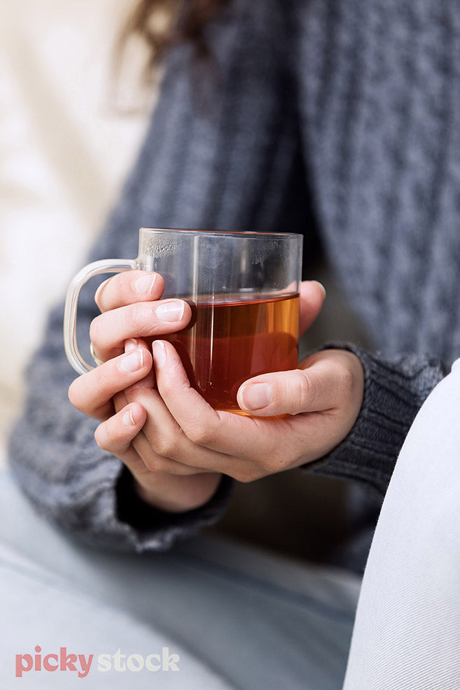 Lady holding glass of herbal tea orange in colour. Body is cropped. Hands in focus with body shallow depth of field. She has brown hair wearing a blue knitted jumper