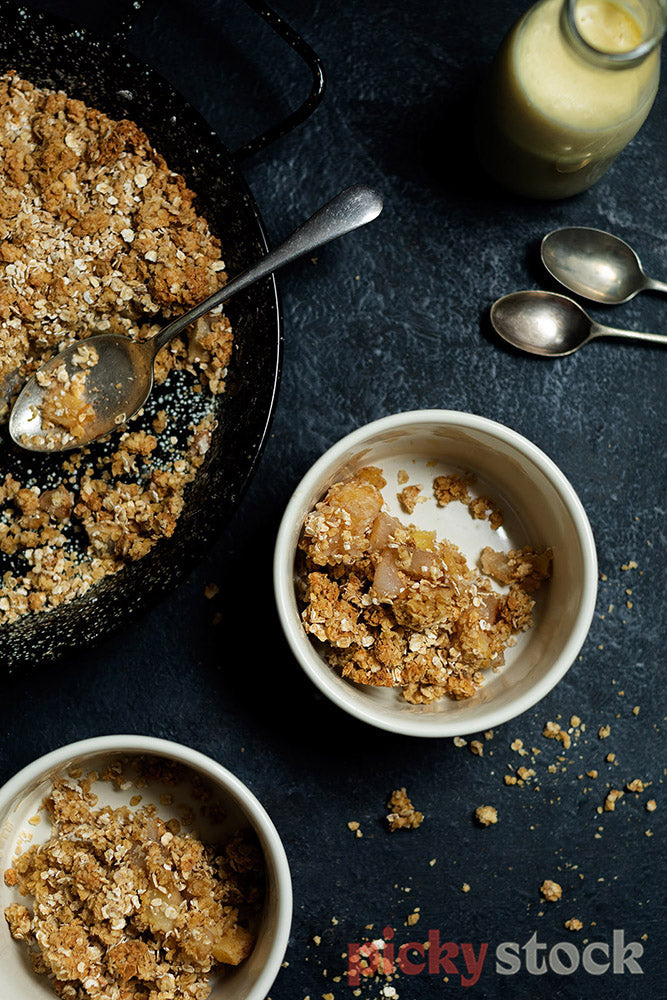 Large dark container of apple crumble with two bowls filled with some. Sprinkles on table. Spoons and glass bottle of custard top of frame.