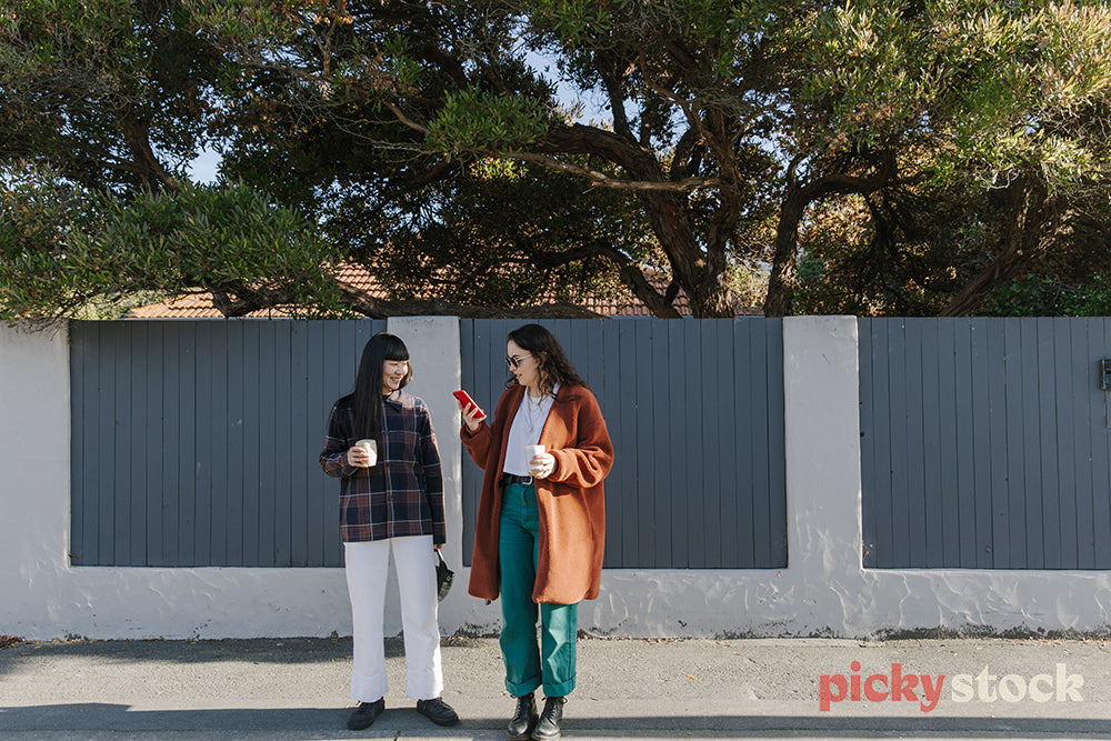 Girl standing on footpath outside home looking at smart phone mobile device. Holding a takeaway coffee in other hand. Wearing long orange jumper with green pants. Friend next to her with coffee cup. White pants dark hair. 
