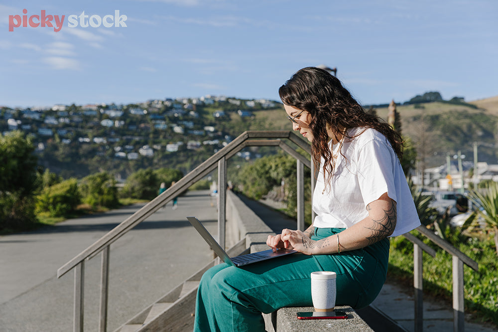 Side view of hip young lady sitting on wall at beach, as she works on laptop.