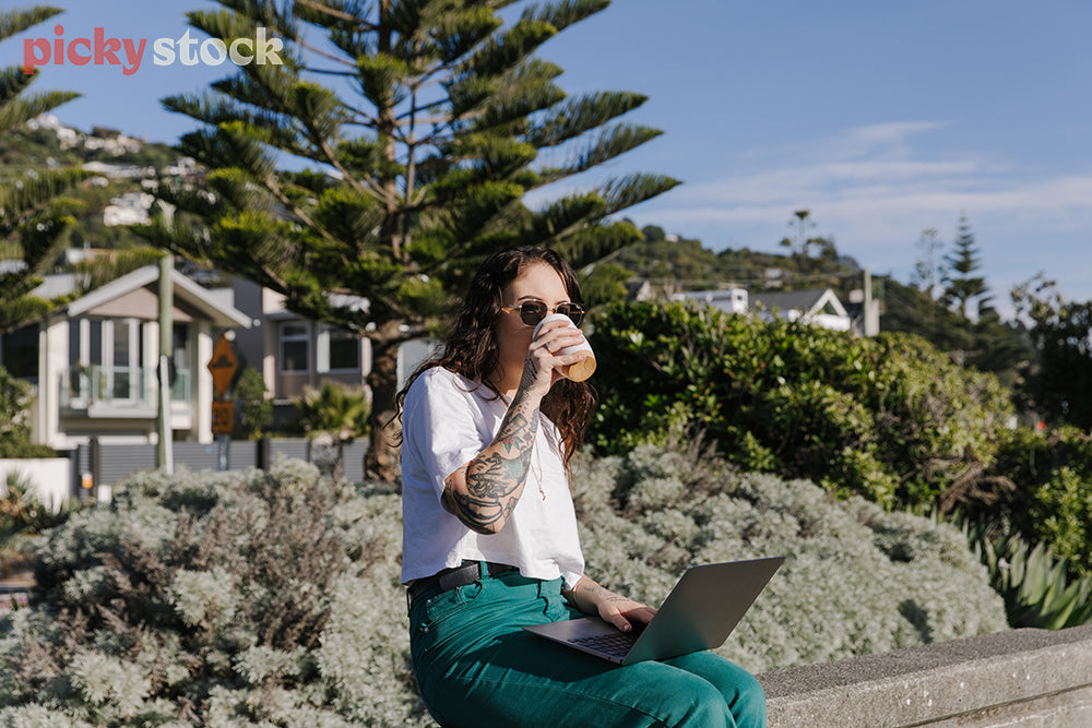 Hip young lady sitting on wall at beach, as she works on laptop on a sunny blue day. 