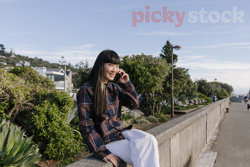 Hip young lady sitting on wall at beach, talking on phone on a sunny blue day. 