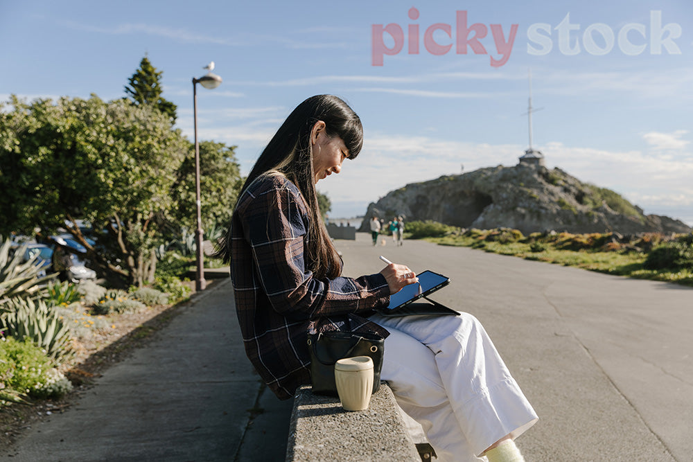 Hip lady sitting on wall at beach, remote working on her tablet with coffee. 