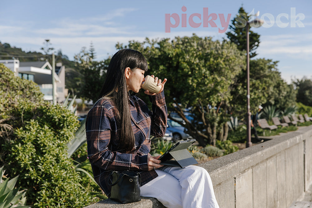 Lady sitting drinking from takeaway coffee mug, holding tablet. Sitting on beach concrete fencing. Trees in back with houses in background. 