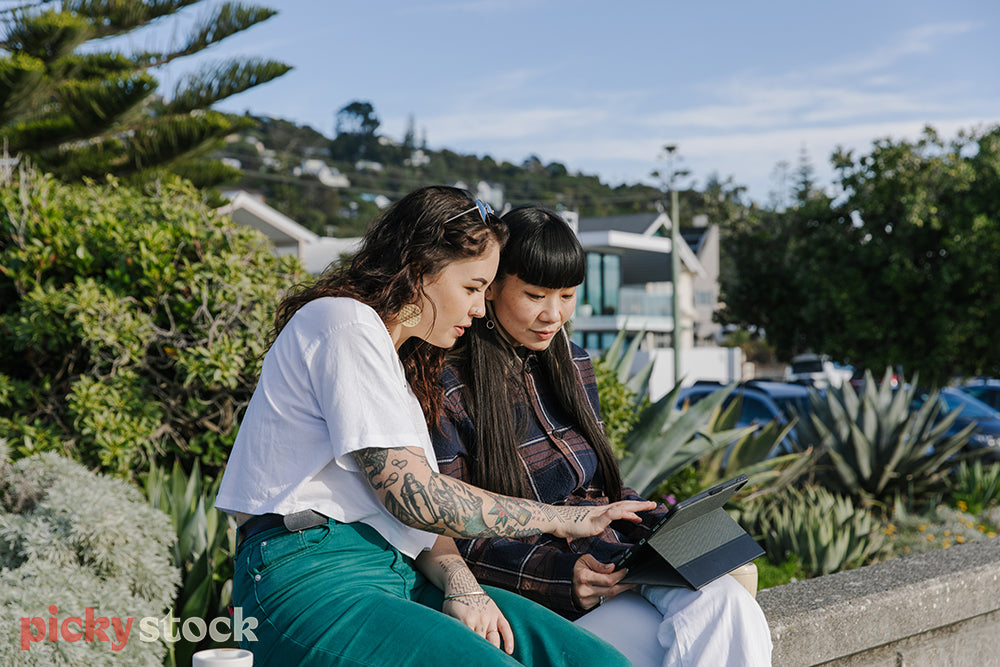 Two wahine sitting along concrete wall at beach, both looking down at a tablet. 