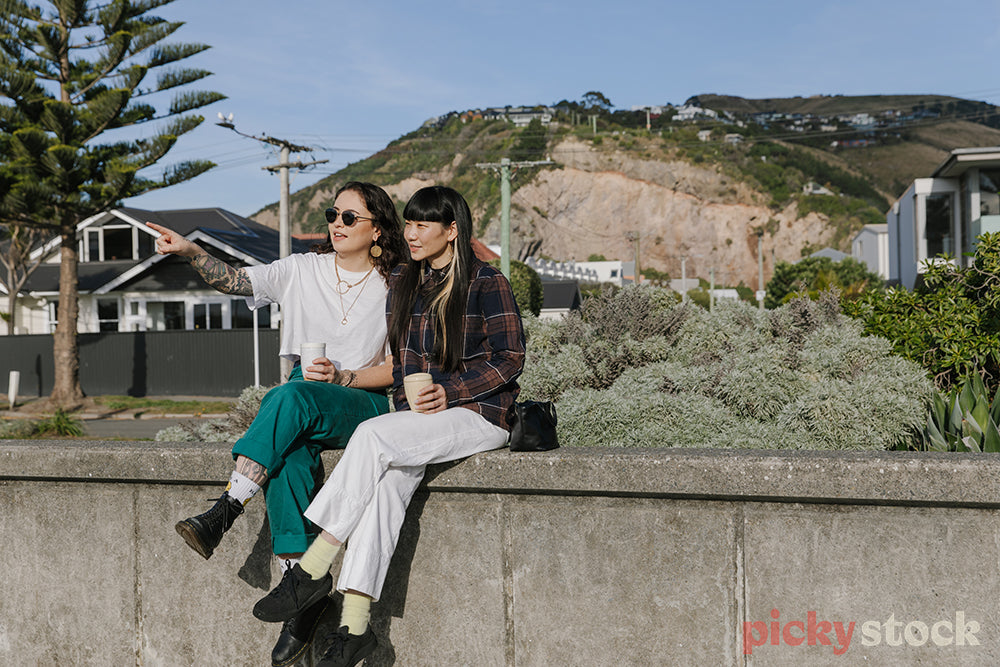Two wahine sitting along concrete wall at beach, with takeaway coffee cups looking out to ocean.