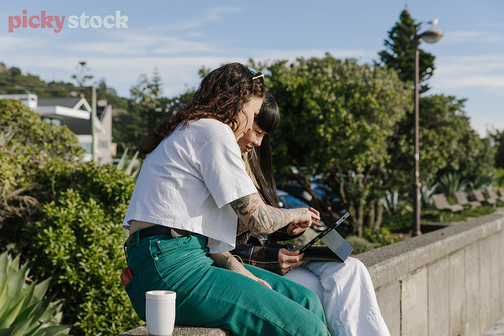 Two wahine sitting along concrete wall at beach, both looking down at a tablet. 