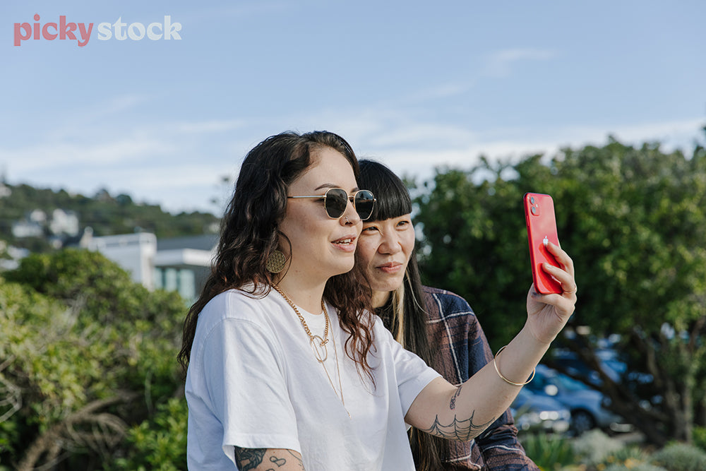 Two friends near waterfront, taking a selfie on red mobile. 