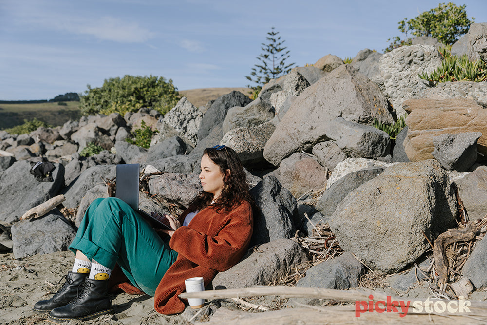 Young wahine in green pants working on laptop sitting against large rocks on beach. 
