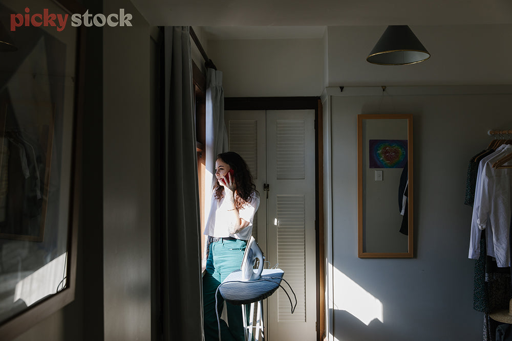 Young wahine standing by bedroom window while talking on mobile phone. Morning light hitting her body causing shadows 