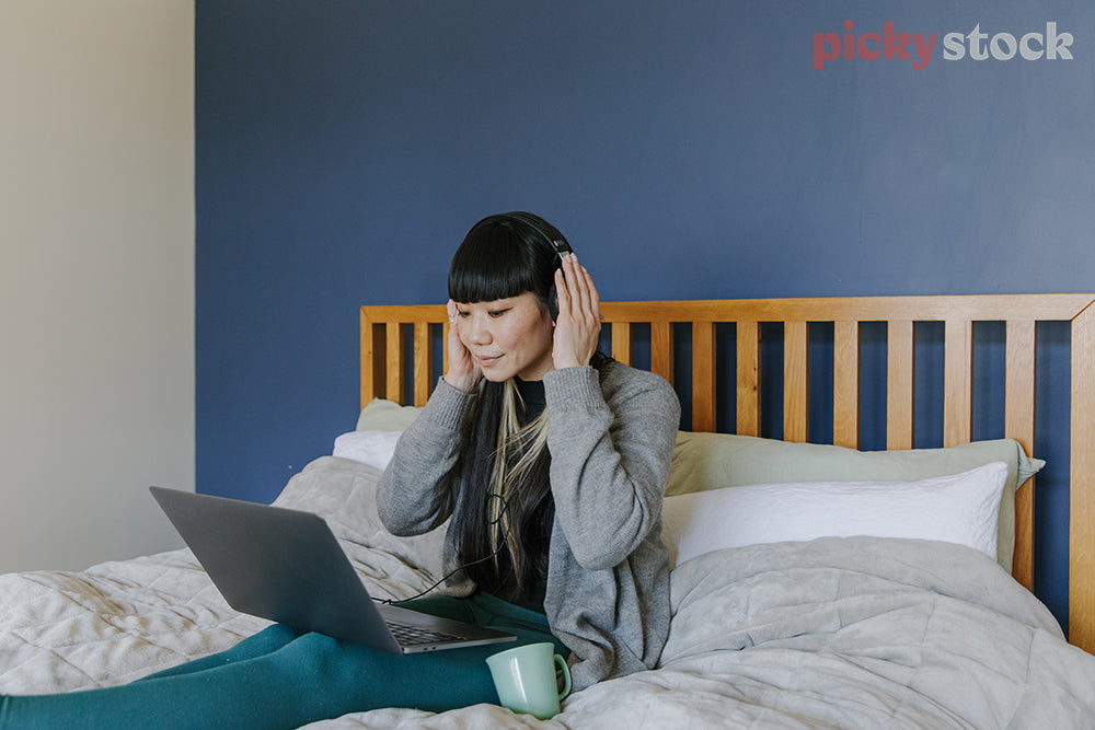 Lady with cup of tea, wearing headphones looking at laptop from bed. Blue background