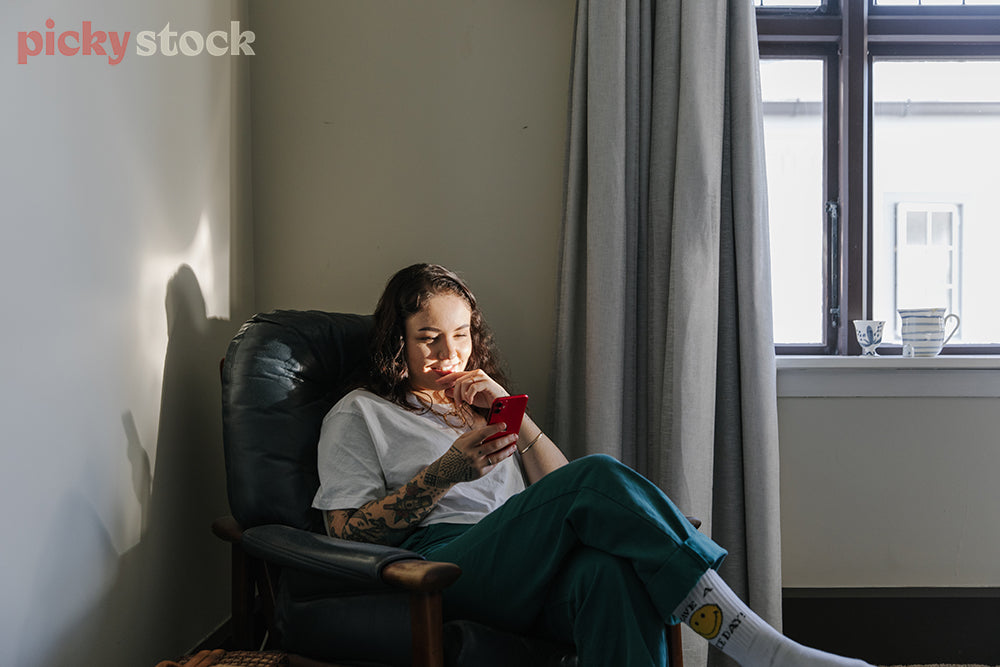Young lady sitting on chair by window. Light from window hitting her face. Sitting on leather chair. Girl is looking at mobile phone with a smile on her face. Wearing socks and green pants. Window has a mug on window sill. Curtains are grey. 