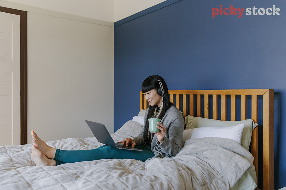lady wearing headphones holding coffee cup, watching a show from her laptop on comfy bed. Large blue wall behind her. 
