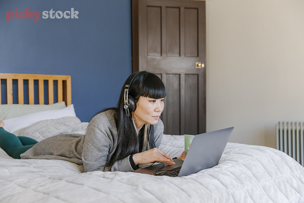 Woman lying on bed wearing headphones looking at laptop.  Blue wall, grey jersey. 