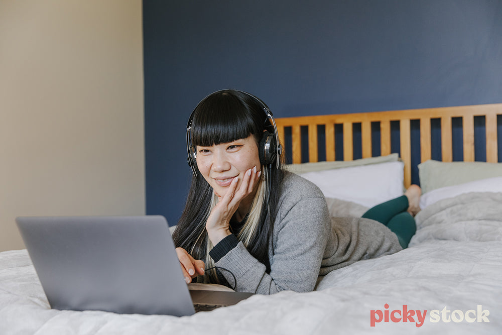 Woman lying on bed wearing headphones watching laptop