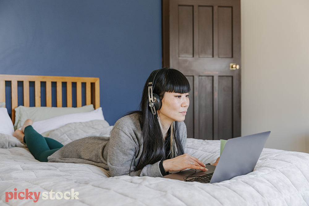 Woman lying on bed, wearing headphones watching laptop screen 
