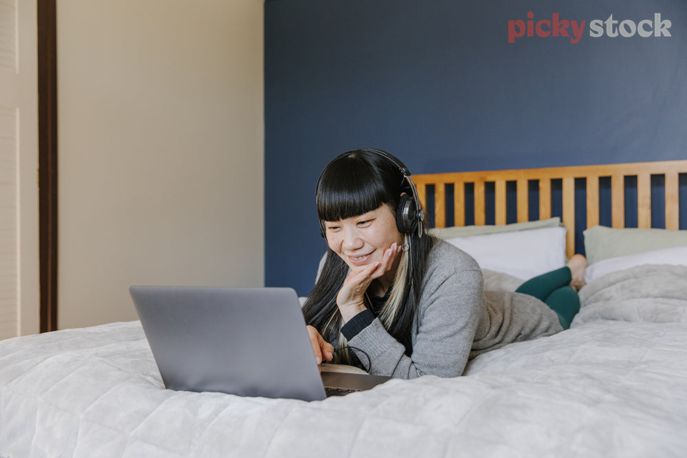 Woman lying on bed, wearing headphones watching laptop screen 