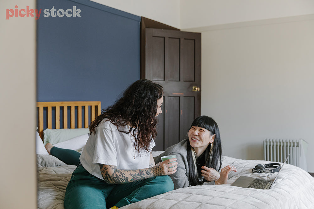 Two female friends sitting on a bed having a laugh, cuppa in hand 