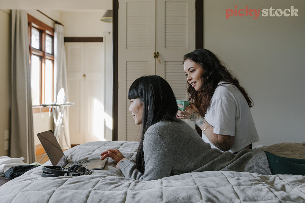 Two female friends sitting on a bed looking at laptop screen in a bedroom. Cupboard and windows in background. 