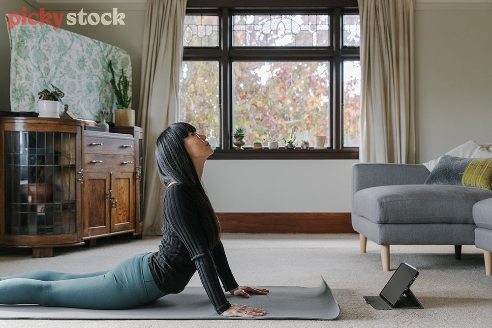 Woman practicing yoga on mat from lounge following along on tablet. Completing the Upward-Facing Dog Pose.