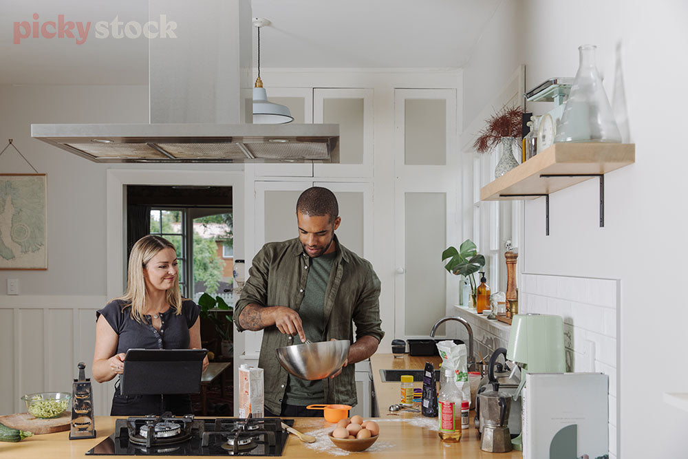 Male and female standing in modern kiwi kitchen following a recipe on a tablet Man holding bowl. 