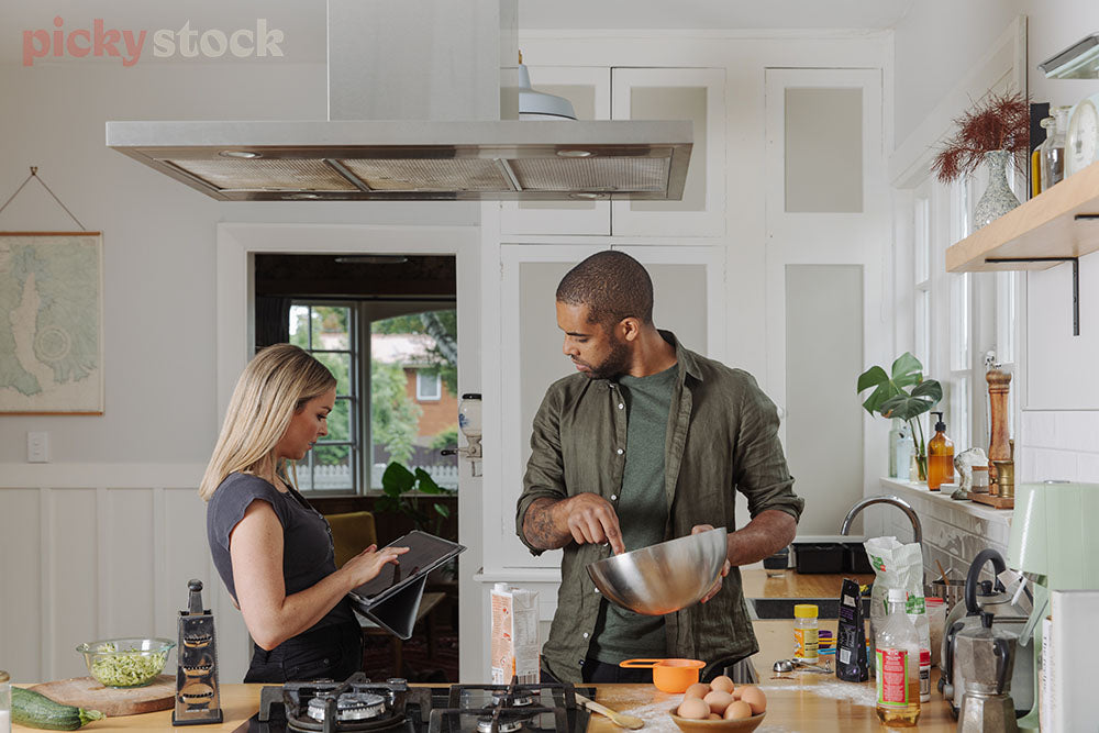 Couple cooking together in kitchen. Following along on a tablet, man holding stainless steel bowl. 