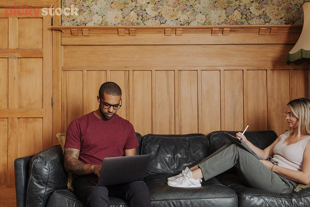 Man wearing glasses sitting on couch wearing red tee shirt working on laptop. Girl sitting across on the couch with legs on the seat. Wearing clear glass frames, white shoes. Background is a wooden wall with wood door. 