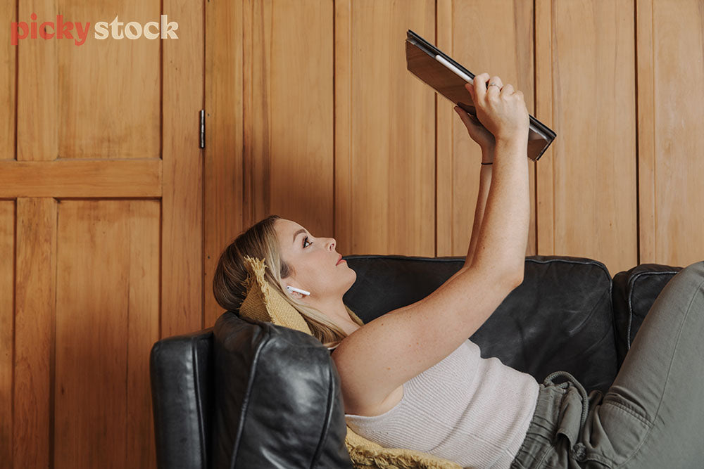 Girl with white ear pods looking up at tablet while she lays against a black leather couch. Wooden walls and door in background. 