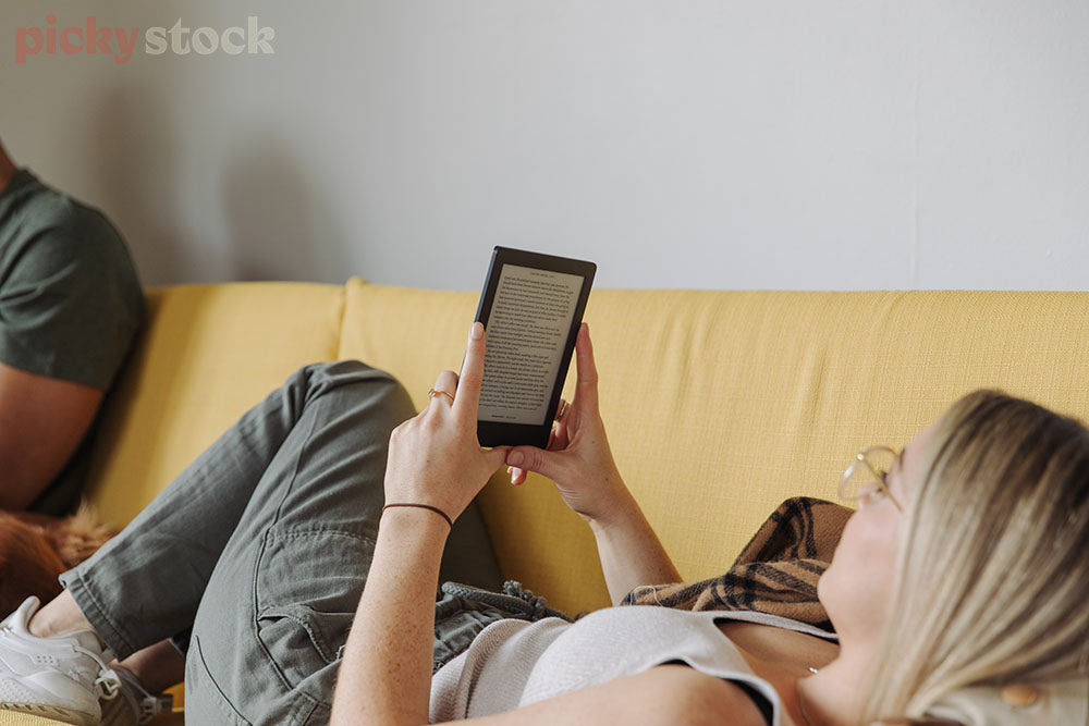 Girl lying on bright yellow couch with blonde hair reading kindle e-book. Outline of male arm left of frame