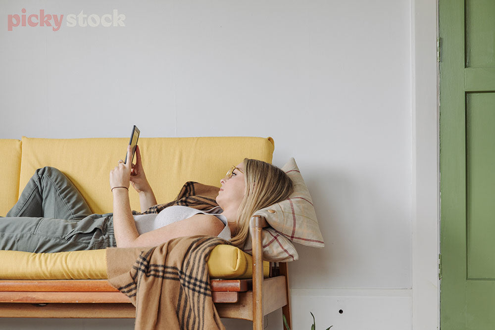 Girl reading kindle on bright yellow couch . Wearing glasses. Green door to right of her. 