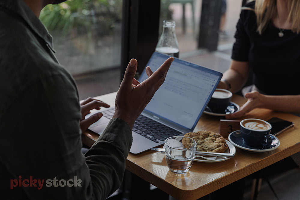 Work meeting at a cafe, focus on laptop screen open with files visible. Coffee and water and cookie on table. Male and female bodies and hands visible cropped at neck.