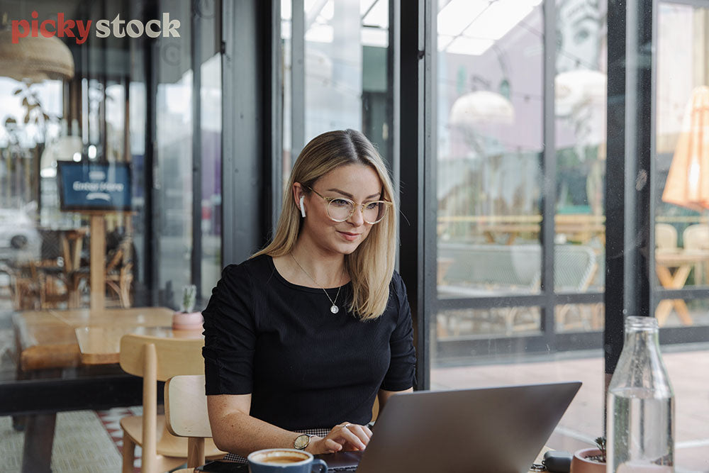 Lady wearing clear glasses and black top sitting at cafe typing on laptop. Coffee next to her. Cafe is modern with large windows. She is wearing a watch 