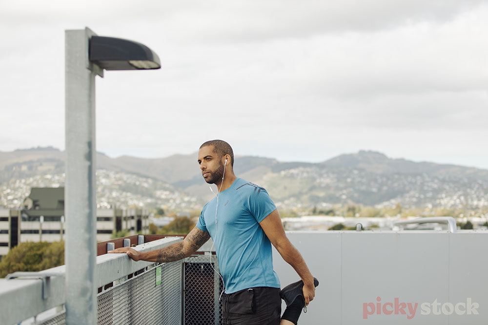 Man wearing headphones wearing a bright blue top stretching before starts a run. Wearing black shoes. View of Christchurch in background. 