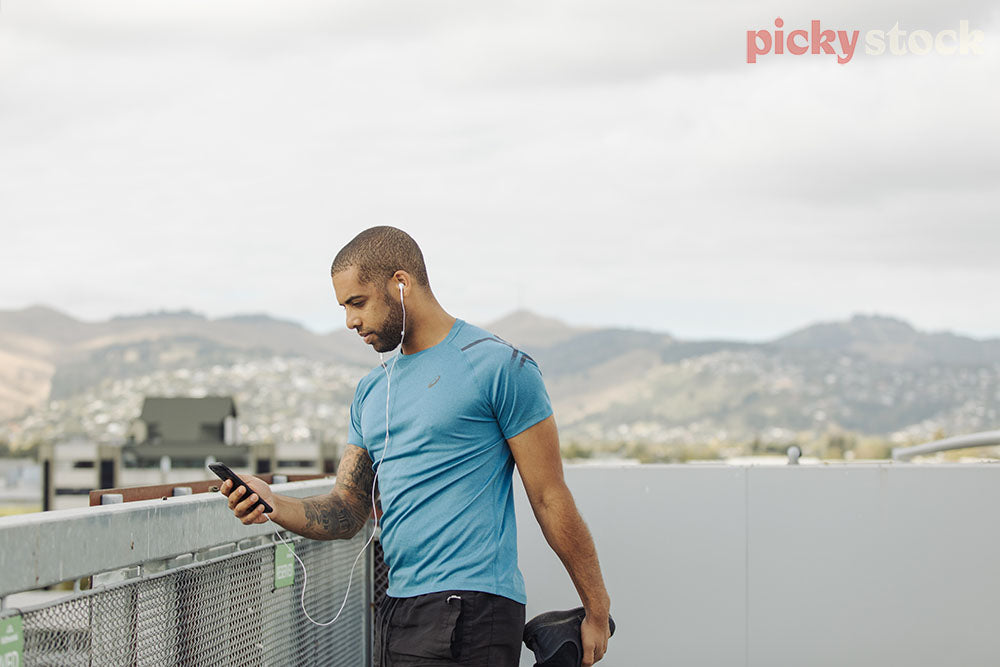 Man wearing headphones checking phone prior to run. Stretching while holding phone in other hand. 