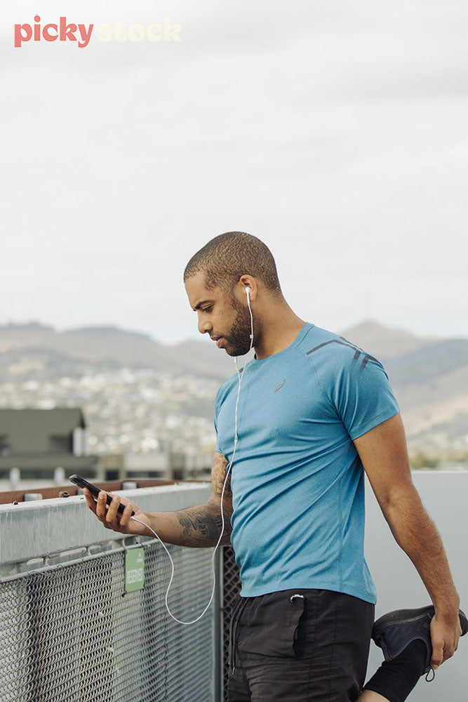 Portrait image of man in blue running tea stretching with one leg and holding mobile device with earphones connected, 