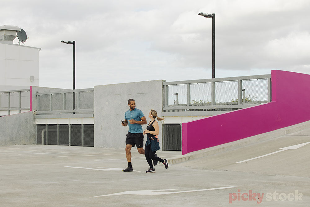 Man in blue shirt and running gear and blonde female in workout clothes running through concrete carpark. Bright magenta pink in background ramp