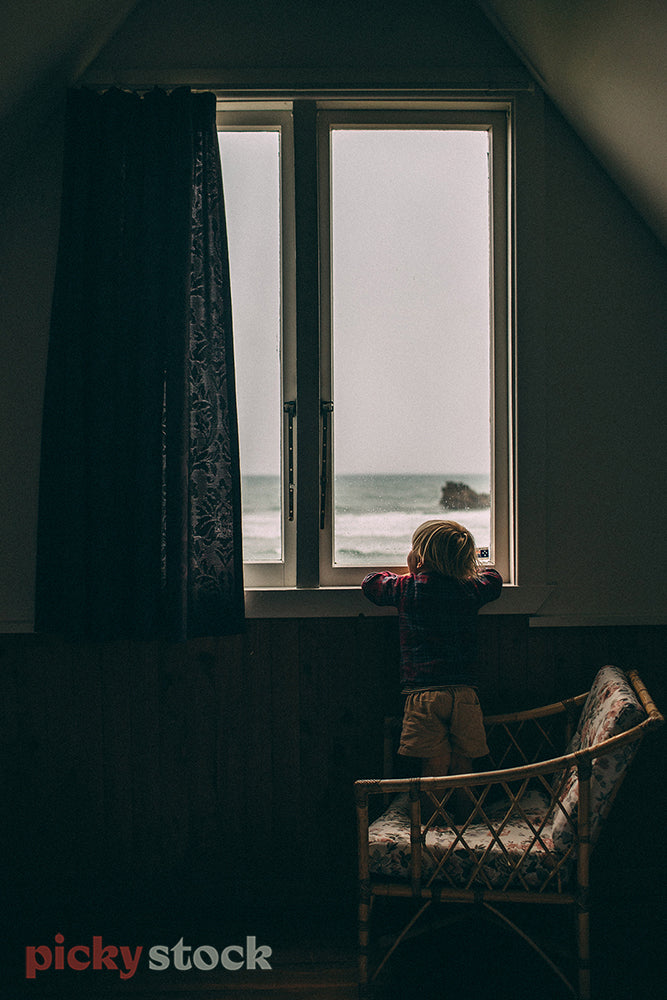 Little boy stands on wicker stool in a dark room out to the sea outside on a grey day
