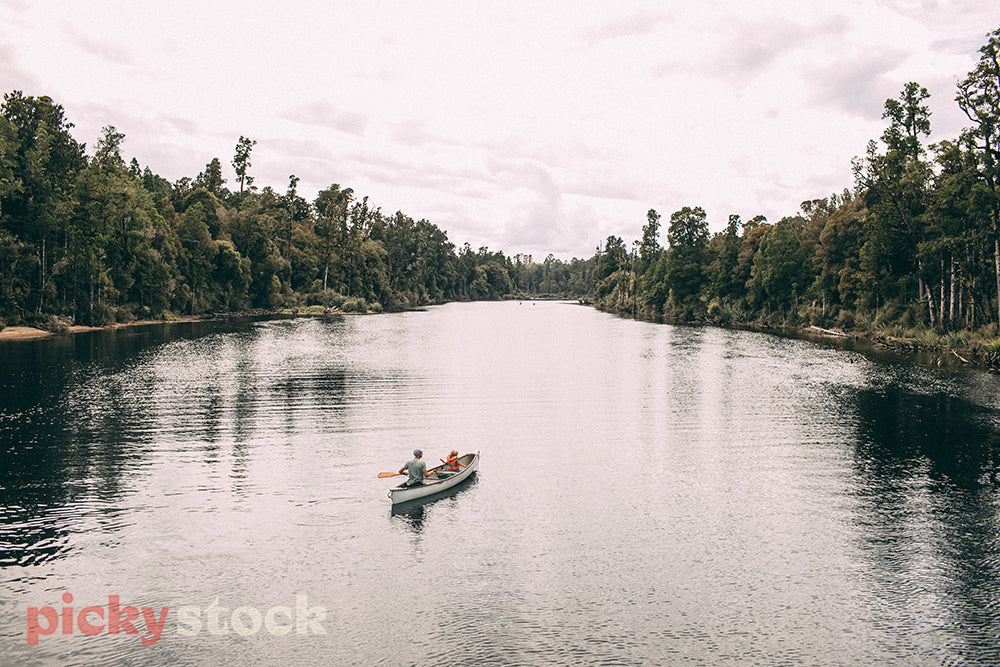 Father and son paddle a canoe on a river surrounded by native New Zealand forest