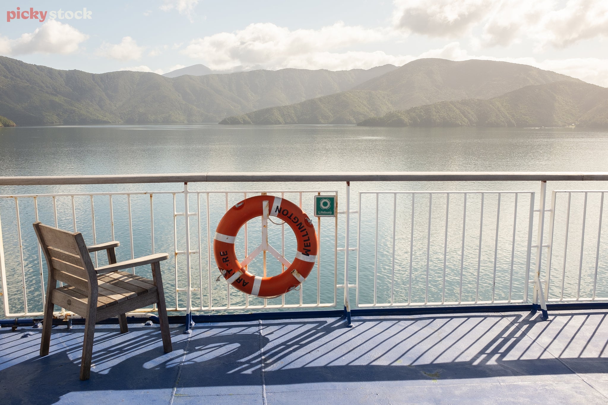 Landscape of close of a lifebuoy and small wooden chair behind a white metal handrail aboard a blue ship deck. The ship cruises past bays of green hills under clear skys.