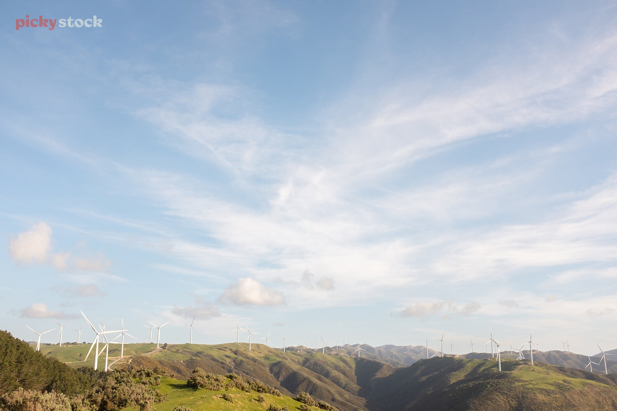 Looking out over a vast wind farm, with turbines peppered between the rolling hills against soft light.