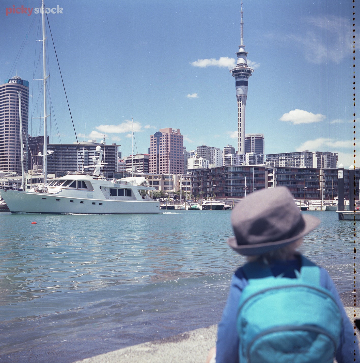 Small child wearing grey hat and blue backpack looking out over Tāmaki Makaurau's waterfront. Auckland waterfront is in view, with Sky Tower middle of frame. Sky is blue with a few clouds around. 