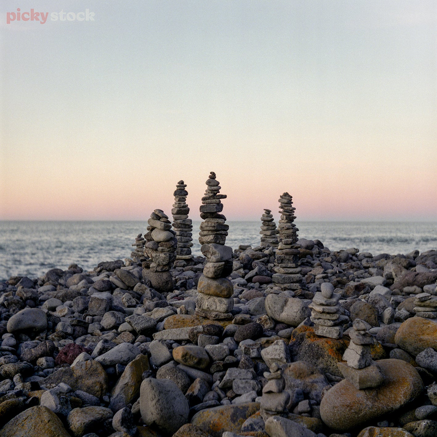 Portrait image of a beach covered in large rocks going out to seat at sunset. Multiple stacks of different sized rocks stacked into towers. 