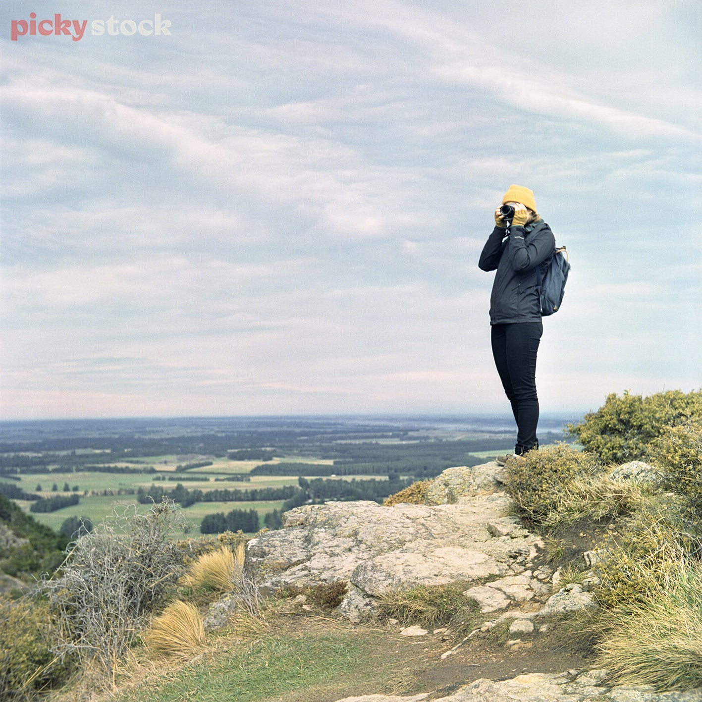 Girl wearina bright yellow beanie and dressed in dark hiking clothes is standing on a rock at the summit of Waspen Falls overlooking the canterbury Plains. Middle of the day, soft blue skies. 