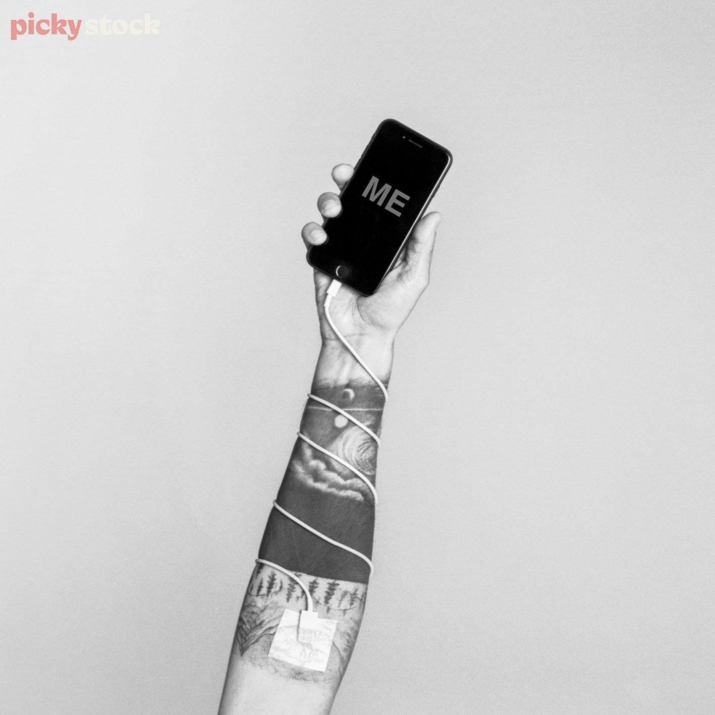 Male hand holding up mobile device with charging cable sellotaped to his arm. On the screen the word ME is written in capslock. Image is black and white. 