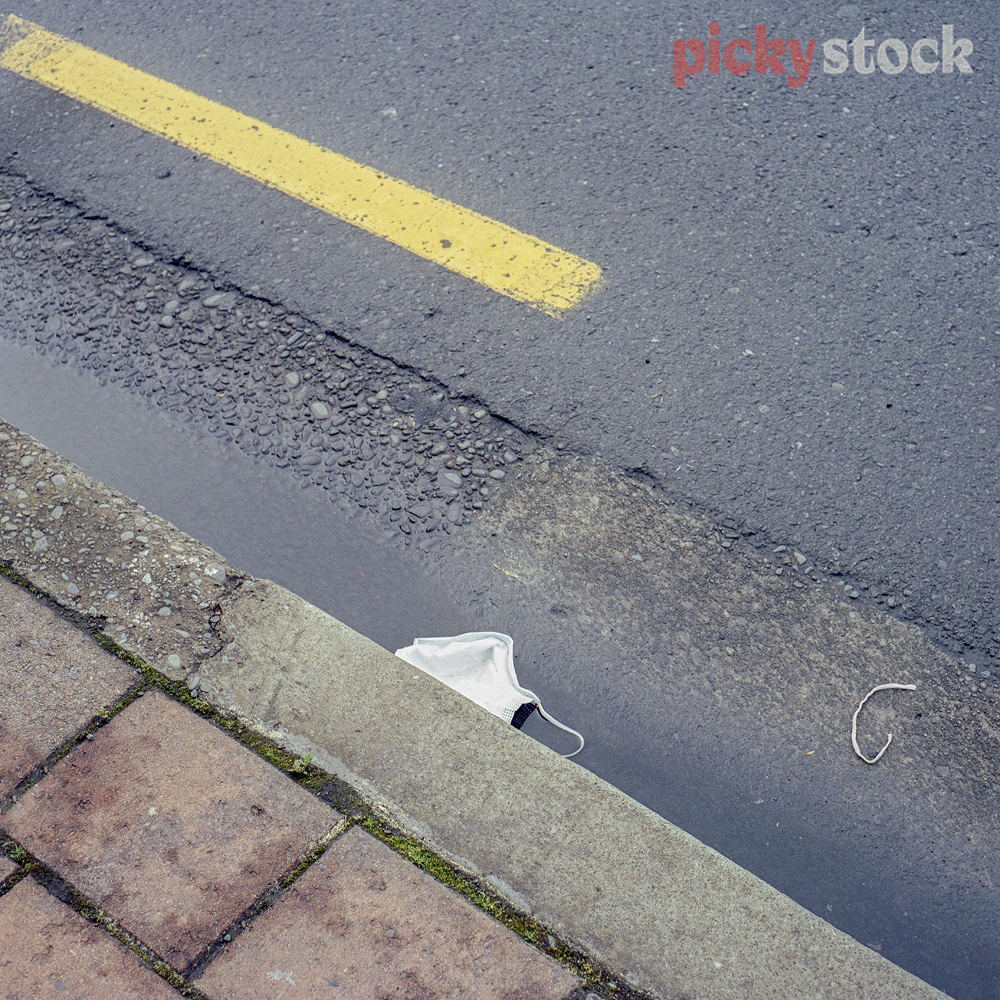 Surgical mask dropped on road. Mask in wet gutter, on road. Yellow road marking line also visible. 