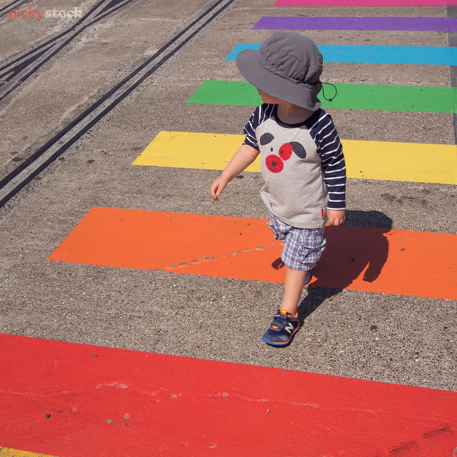 Little boy walks across colourful rainbow road markings, pedestrian crossing. Boy is wearing a dark bucket hat. 