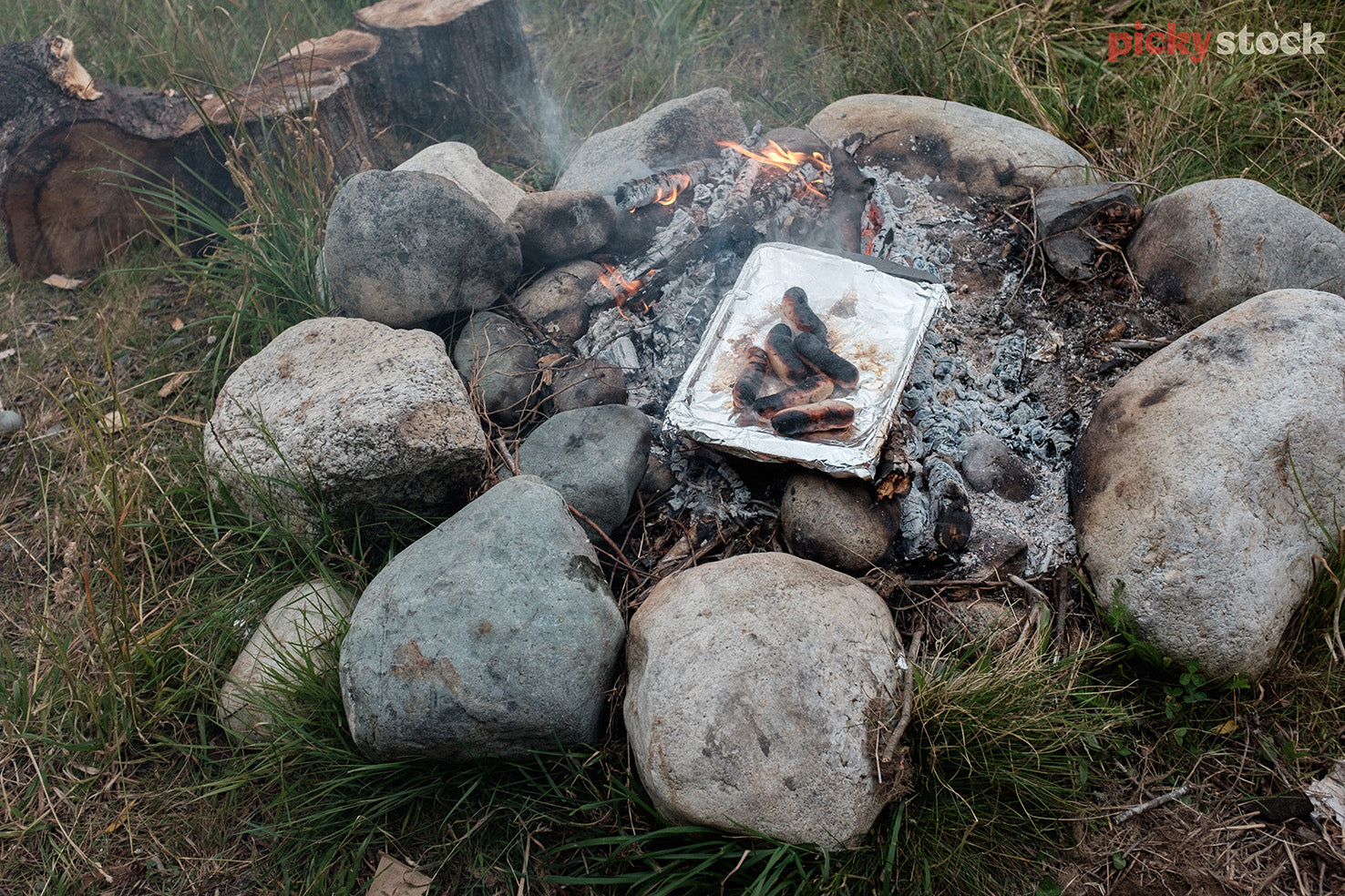 Sausages being cooked on an open outdoor fire. Sitting on a tinfoil tray. Smoke and hot amber flames visible. Large rocks surrouding the outside on grass. 