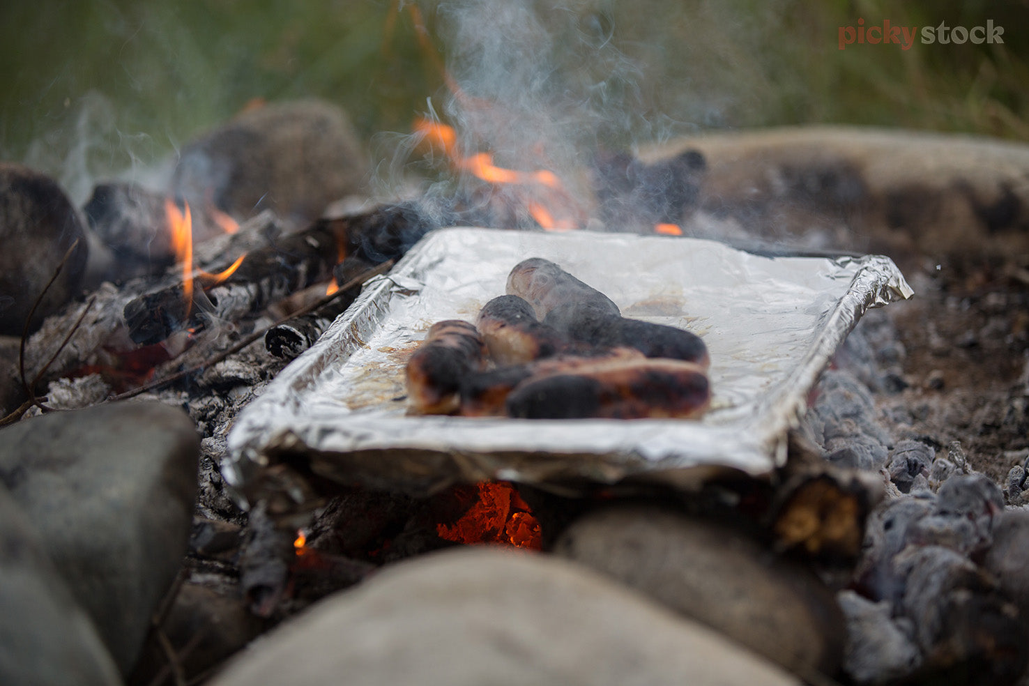 Sausages being cooked on an open outdoor fire. Sitting on a tinfoil tray. Smoke and hot amber flames visible. 